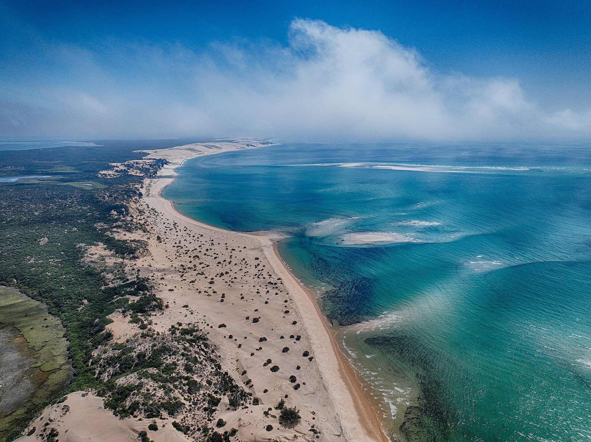 Vista aérea de uma faixa de areia com dunas e vegetação entre mata e lagoa à esquerda e mar azul-turquesa à direita