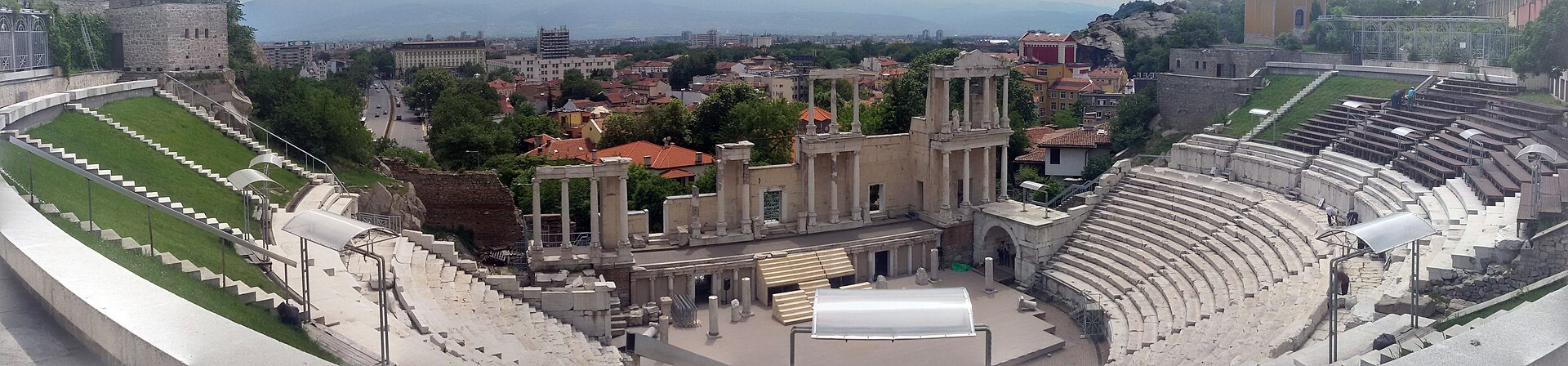 Teatro Romano de Plovdiv em foto panorâmica, com arquibancadas de pedra, colunas da fachada do palco e telhados vermelhos da cidade ao fundo