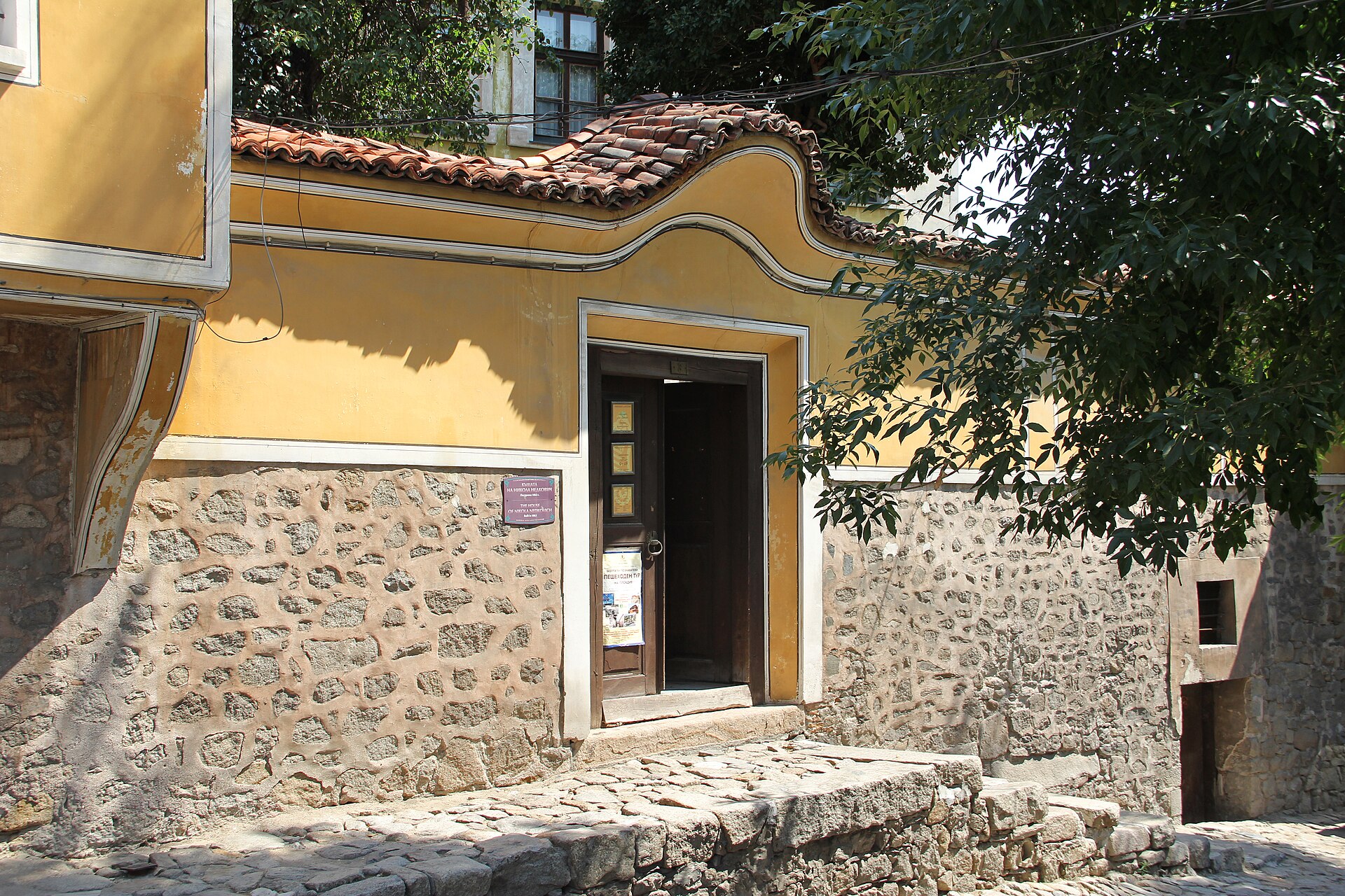 Casa amarela com portal de madeira aberto, telhado de telhas e base de pedra, em uma rua de paralelepípedos da Cidade Velha de Plovdiv