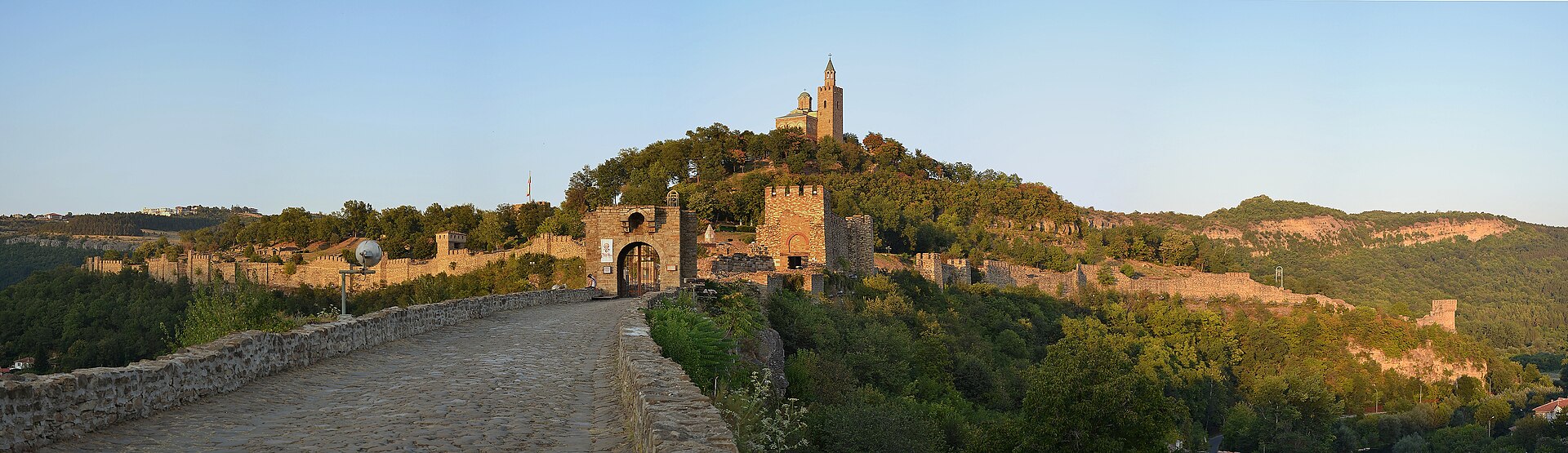 Panorama da fortaleza de Tsarevets ao entardecer, com muralha de pedra, portão em arco, torre de vigia e a igreja de campanário no topo da colina arborizada