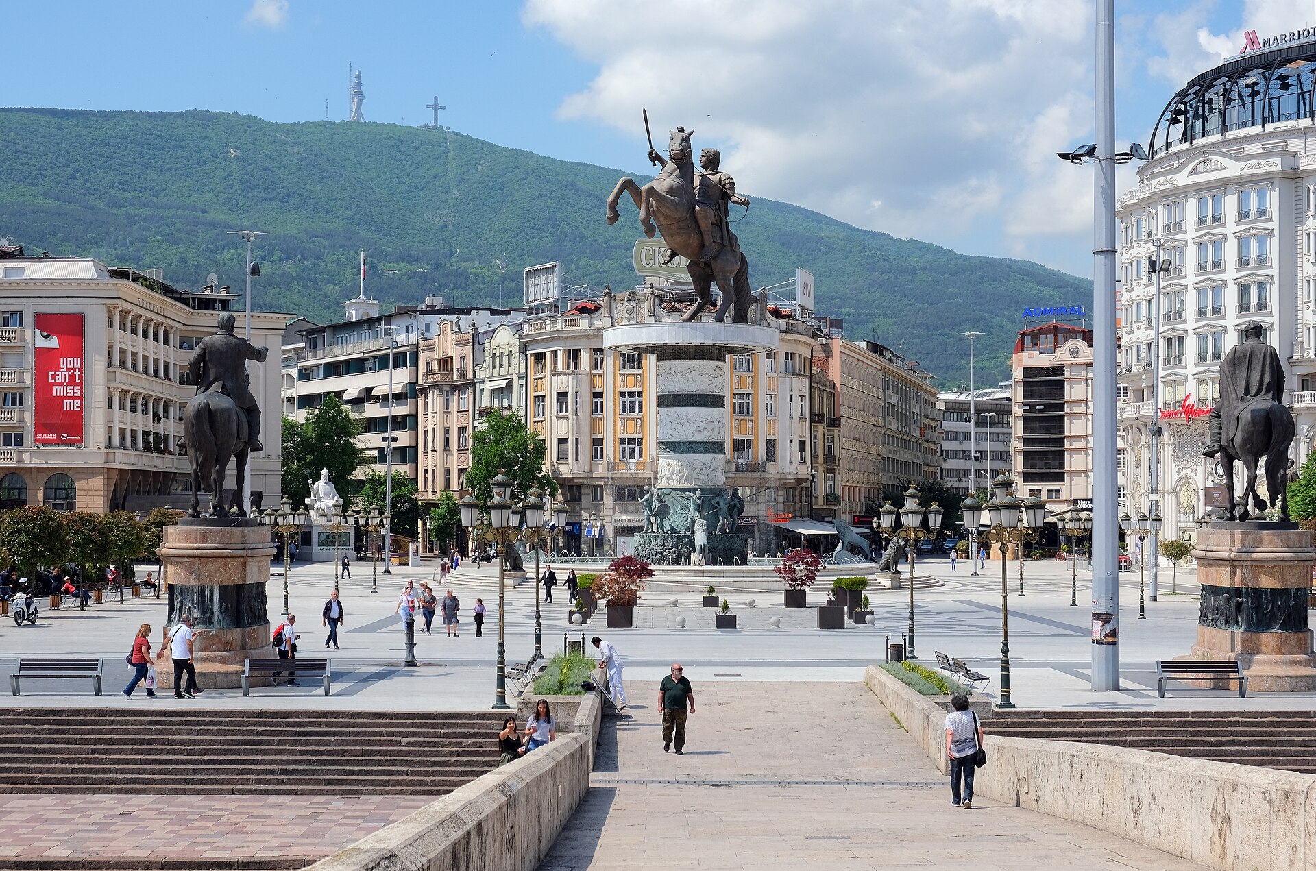 Praça Macedônia, em Skopje, vista da Ponte de Pedra, com a estátua equestre de Alexandre, o Grande, sobre a fonte, edifícios claros e uma montanha com cruz ao fundo