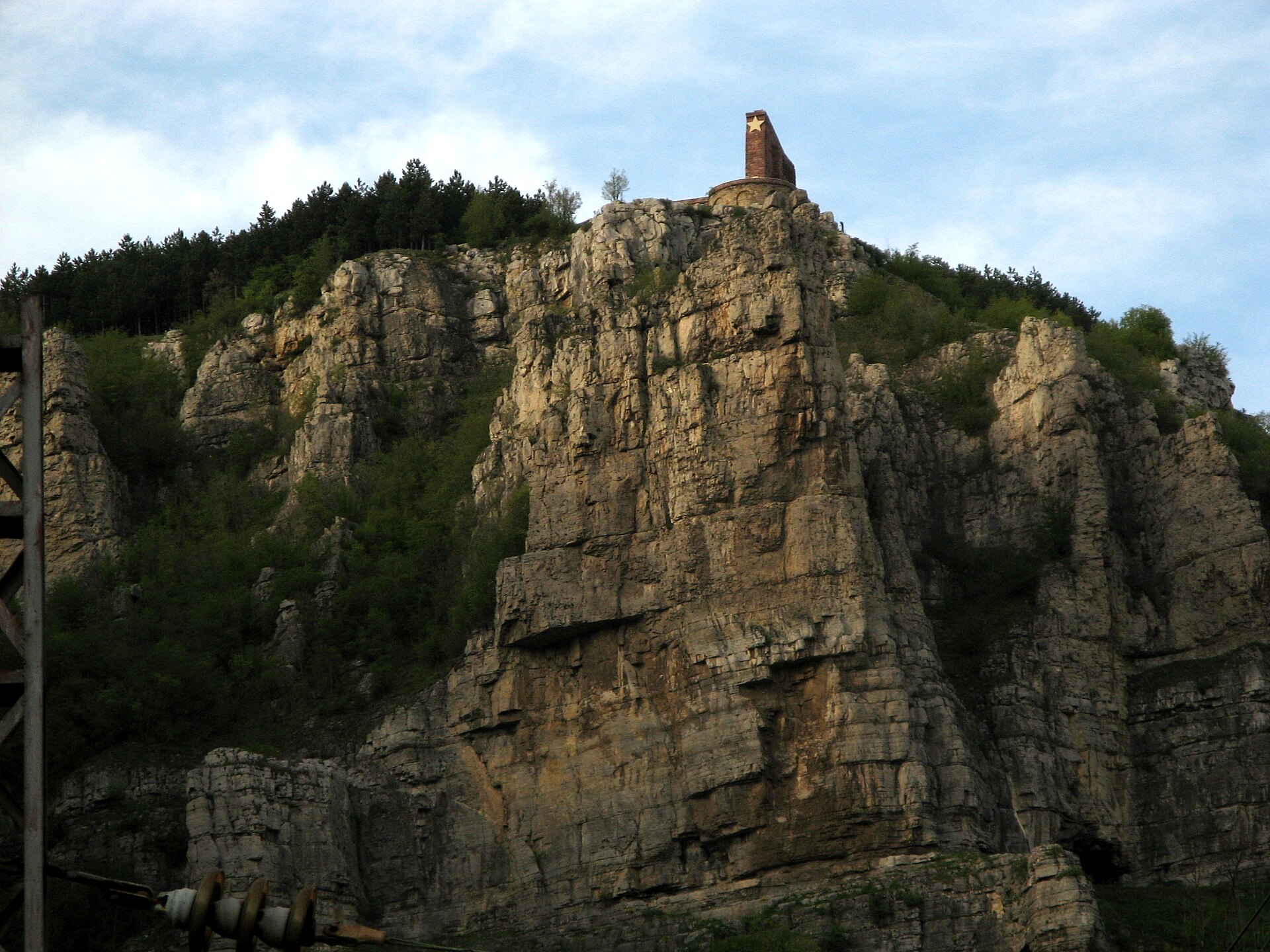 Paredão de calcário nas Rochas de Lakatnik, no Desfiladeiro do Iskar, com um monumento de tijolo com estrela no topo e pinheiros na encosta