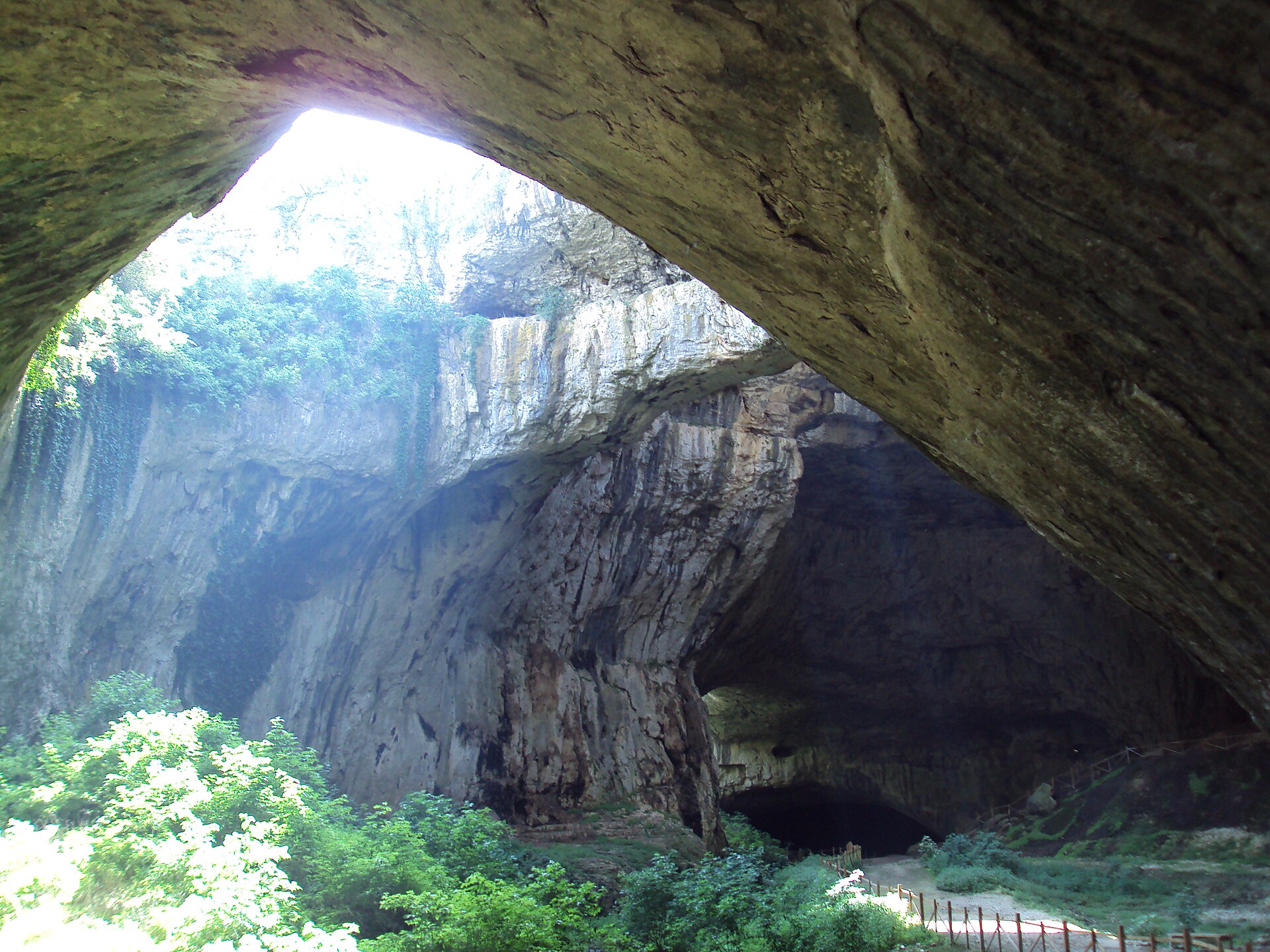 Entrada ampla da Gruta de Devetaki, com arco de rocha, vegetação verde e uma passarela de madeira ao fundo