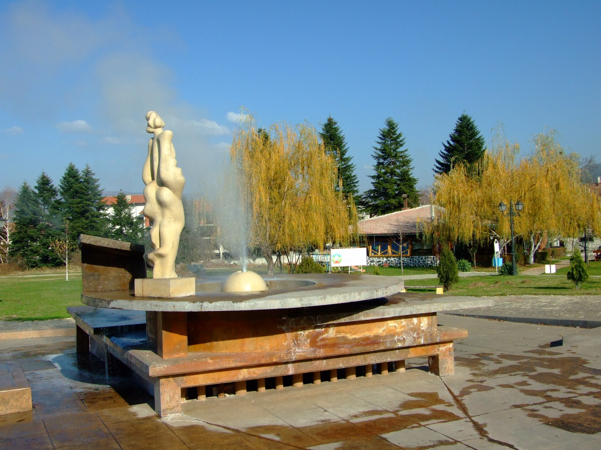 Fonte com escultura clara e jato de água quente com vapor num parque de Sapareva Banya, com salgueiros amarelados e pinheiros ao fundo