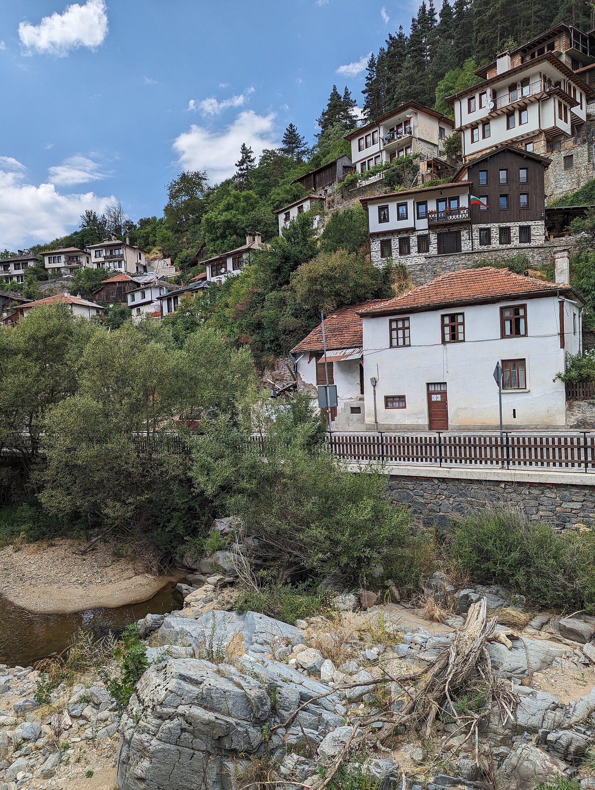 Casas brancas de telhado de telha e fachadas de madeira escura em degraus na encosta arborizada de Shiroka Laka, acima de um rio de leito pedregoso