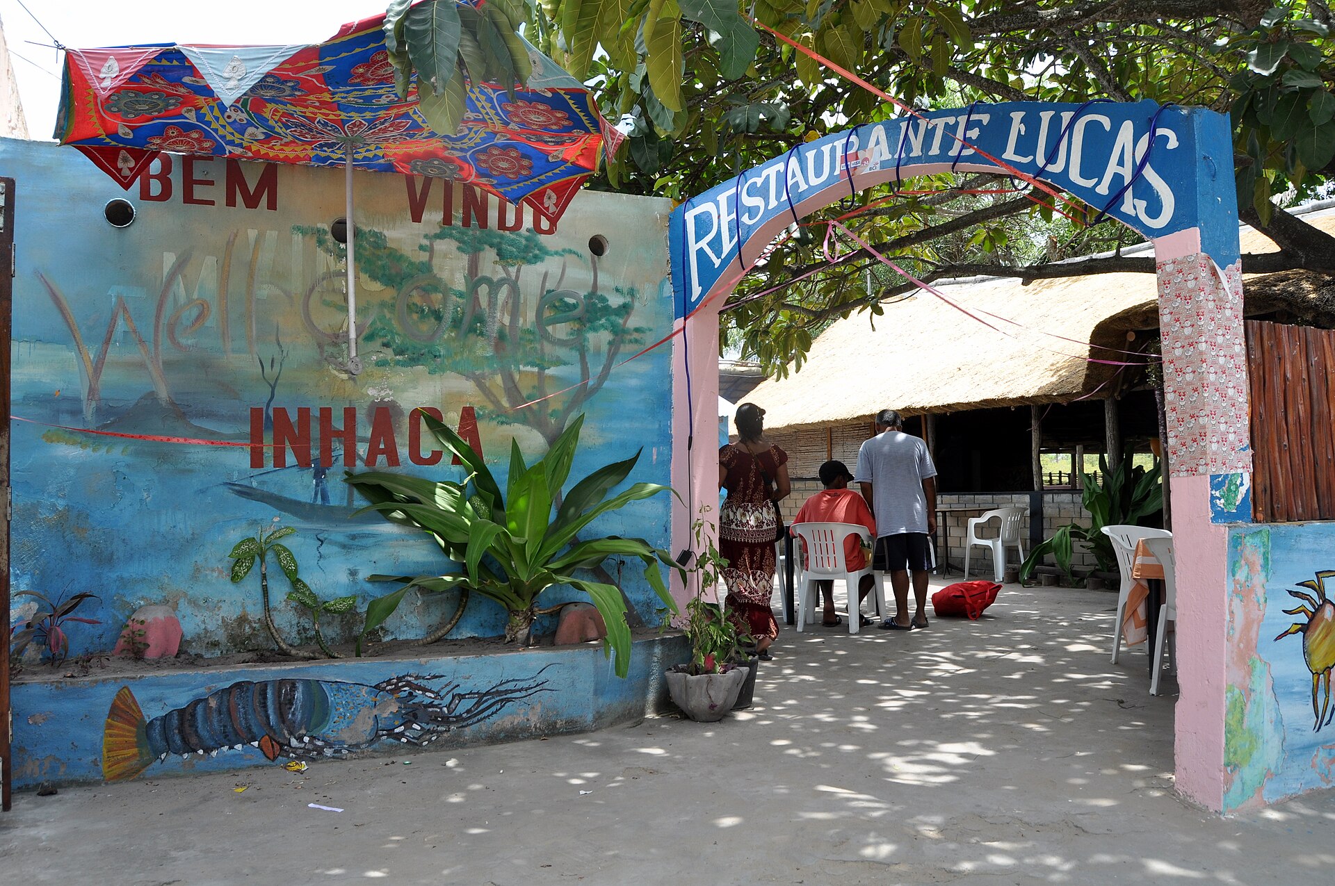 Entrada do Restaurante Lucas, com arco azul e rosa, muro pintado com a mensagem "Bem-vindo Inhaca" e um lagostim, e pessoas ao lado de mesas sob um telhado de palha