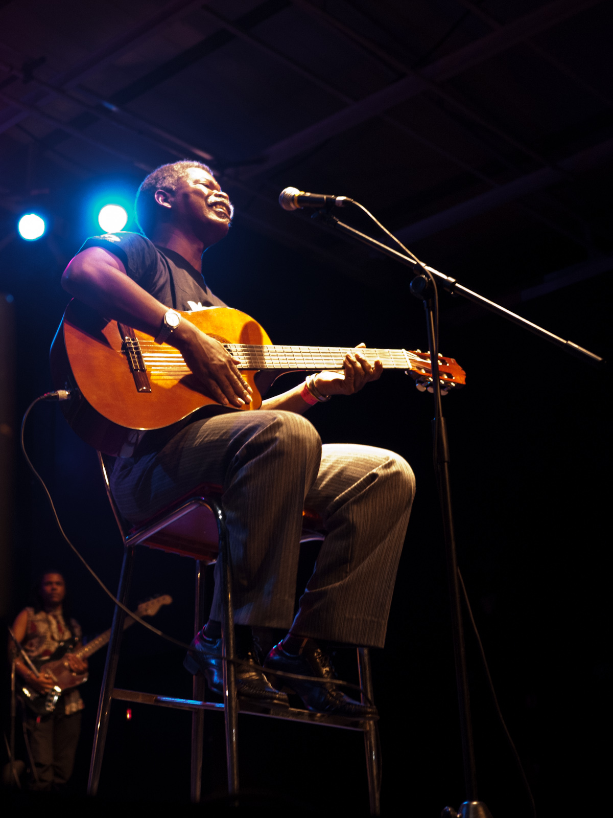 Músico sentado num banquinho cantando e tocando violão no palco, diante de um microfone, sob luzes azuis, com um guitarrista ao fundo