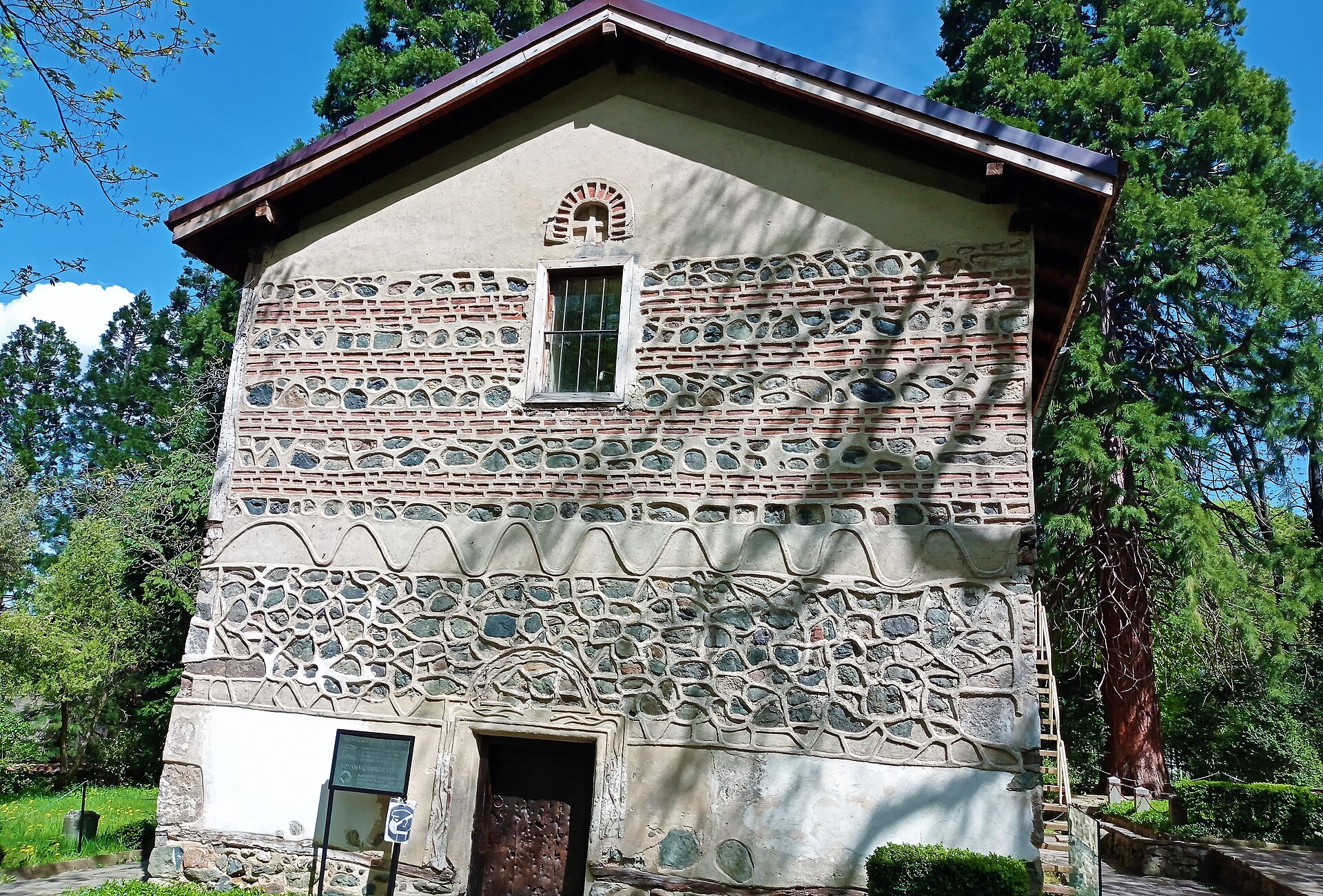 Fachada de pedra e tijolo da pequena Igreja de Boyana, com porta de madeira e árvores altas ao redor