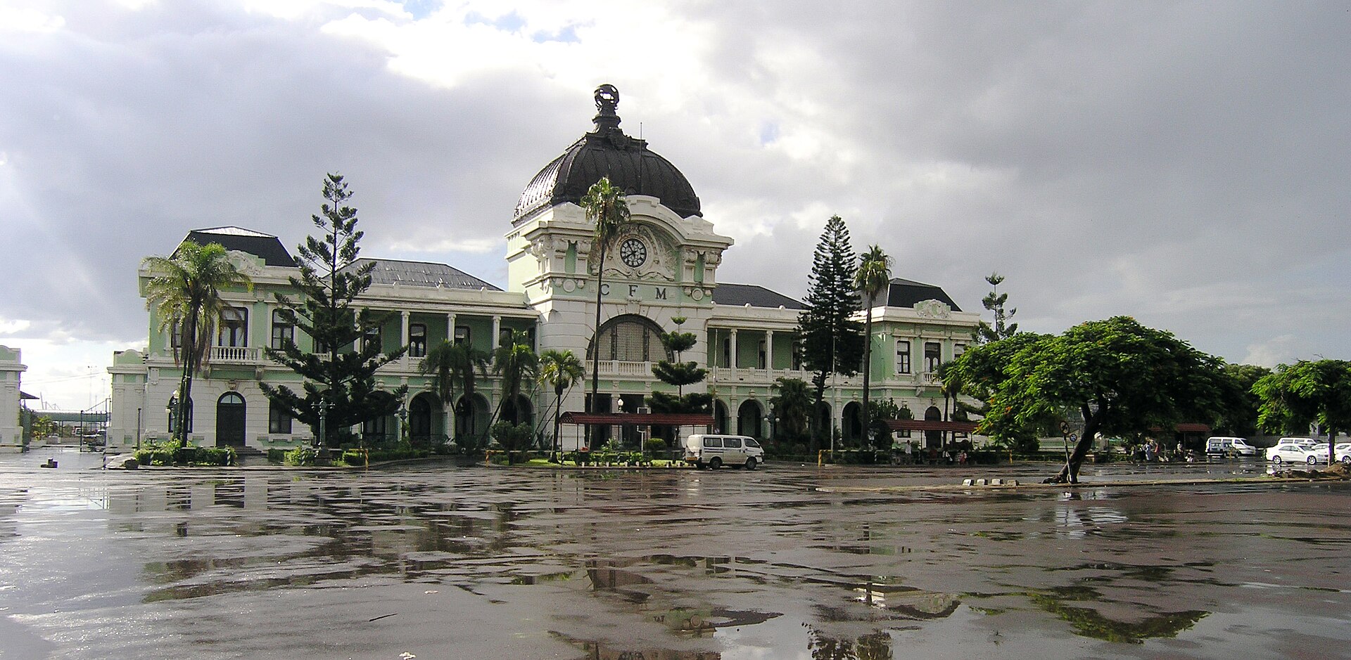 Fachada verde-clara e branca da Estação Central de Maputo, com cúpula escura, relógio e as letras CFM, e o pátio molhado de chuva à frente