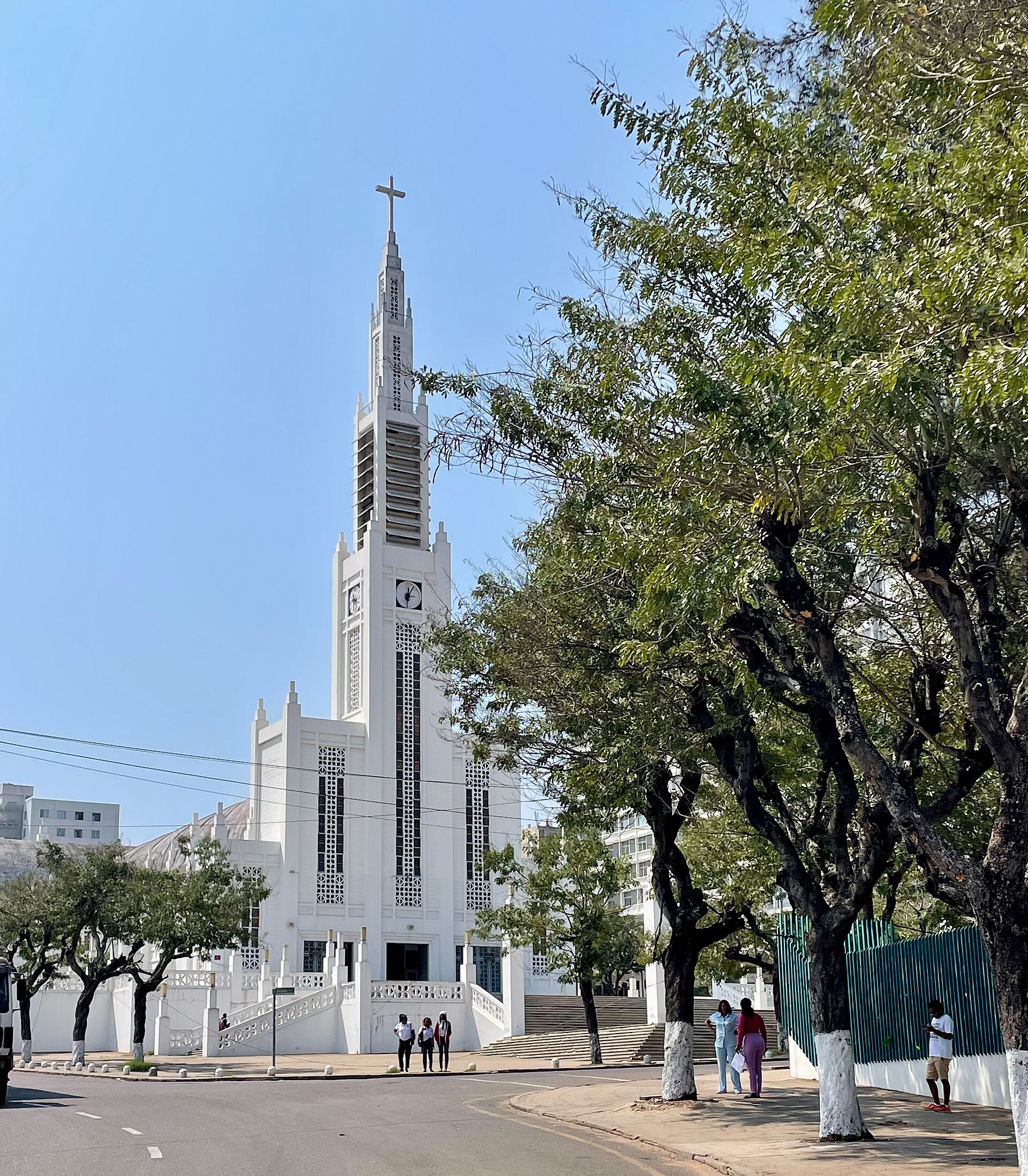 Catedral branca de torre alta com cruz no topo e fachada de painéis vazados, vista da rua entre árvores, com algumas pessoas na calçada