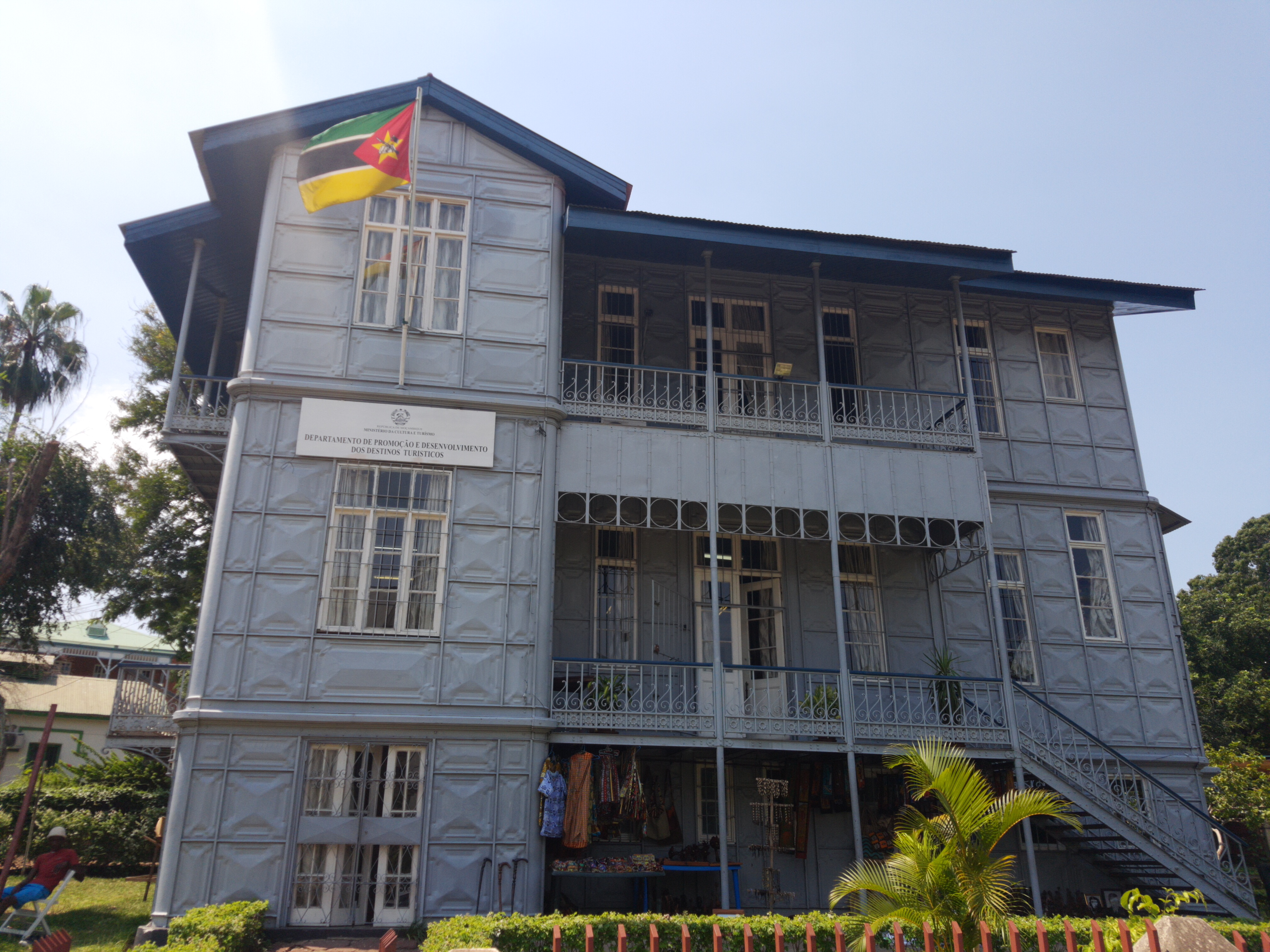 Casa de Ferro de Maputo, prédio de painéis metálicos cinza com varandas de grade trabalhada, escada externa, bandeira de Moçambique e barraca de tecidos no térreo