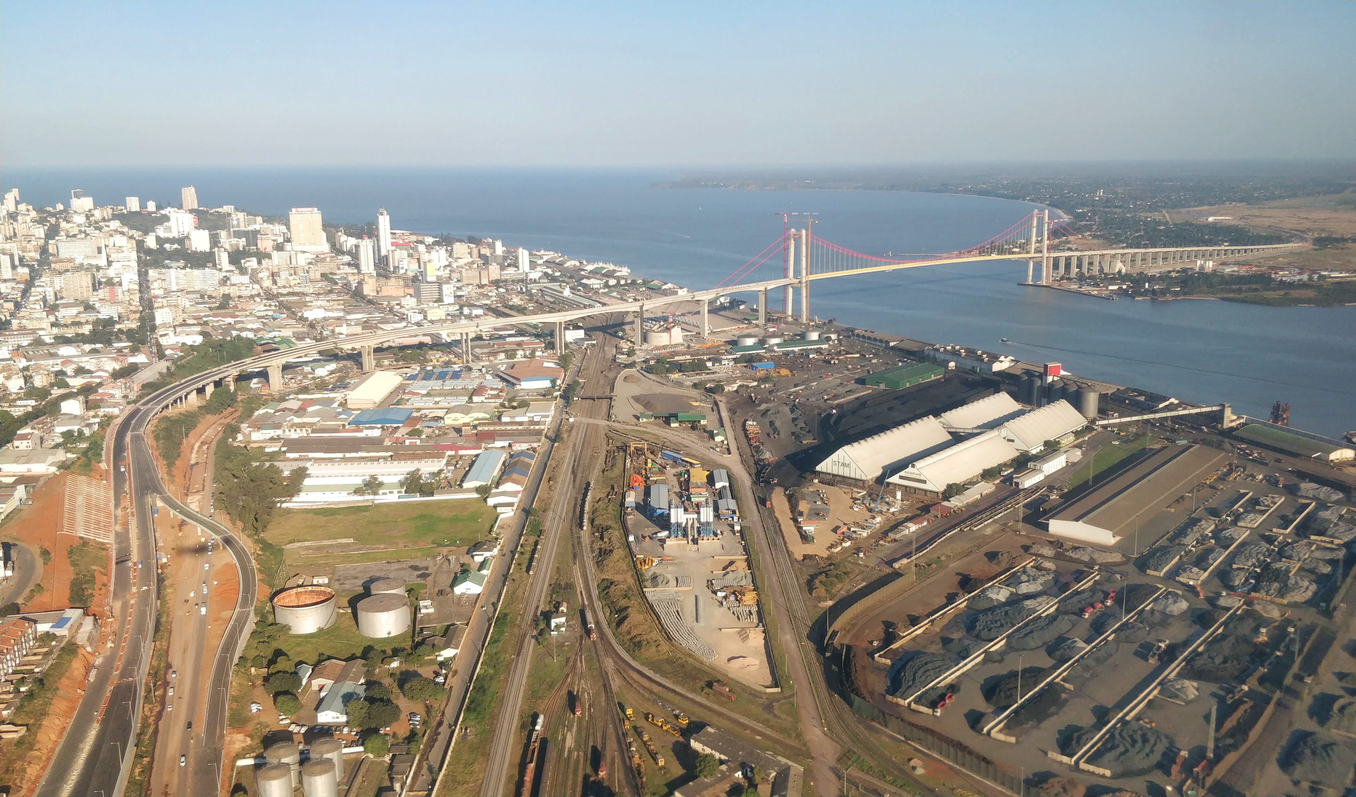 Vista aérea da orla de Maputo com os prédios do centro, o porto com pátios de carga e a ponte suspensa Maputo-Katembe de cabos vermelhos sobre a baía