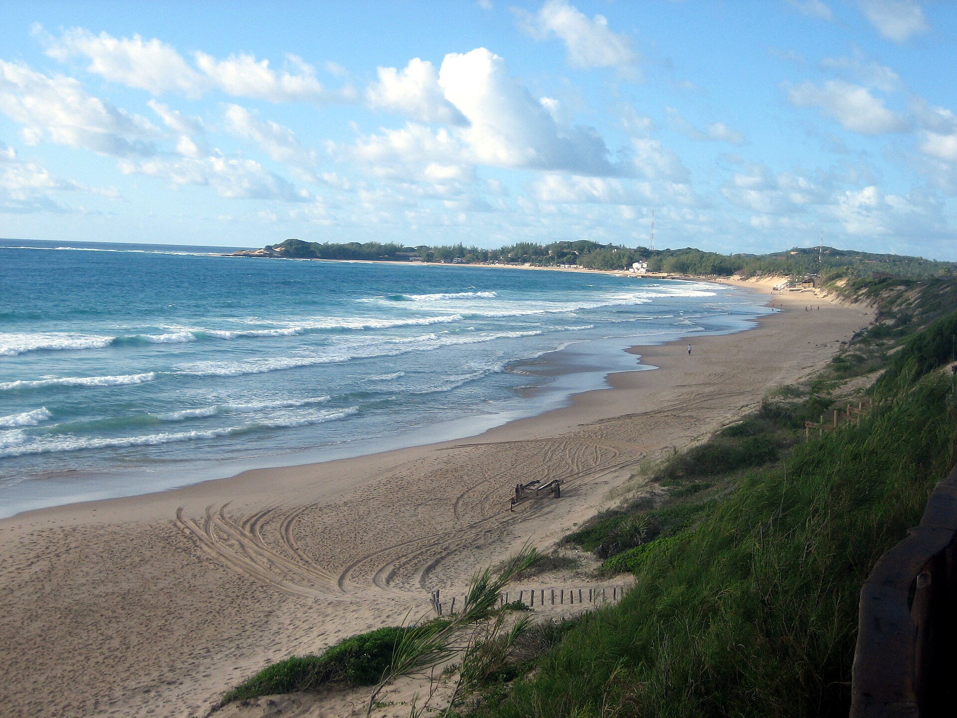 Praia larga de areia clara com ondas quebrando à esquerda, dunas cobertas de vegetação à direita e uma ponta de mata ao fundo