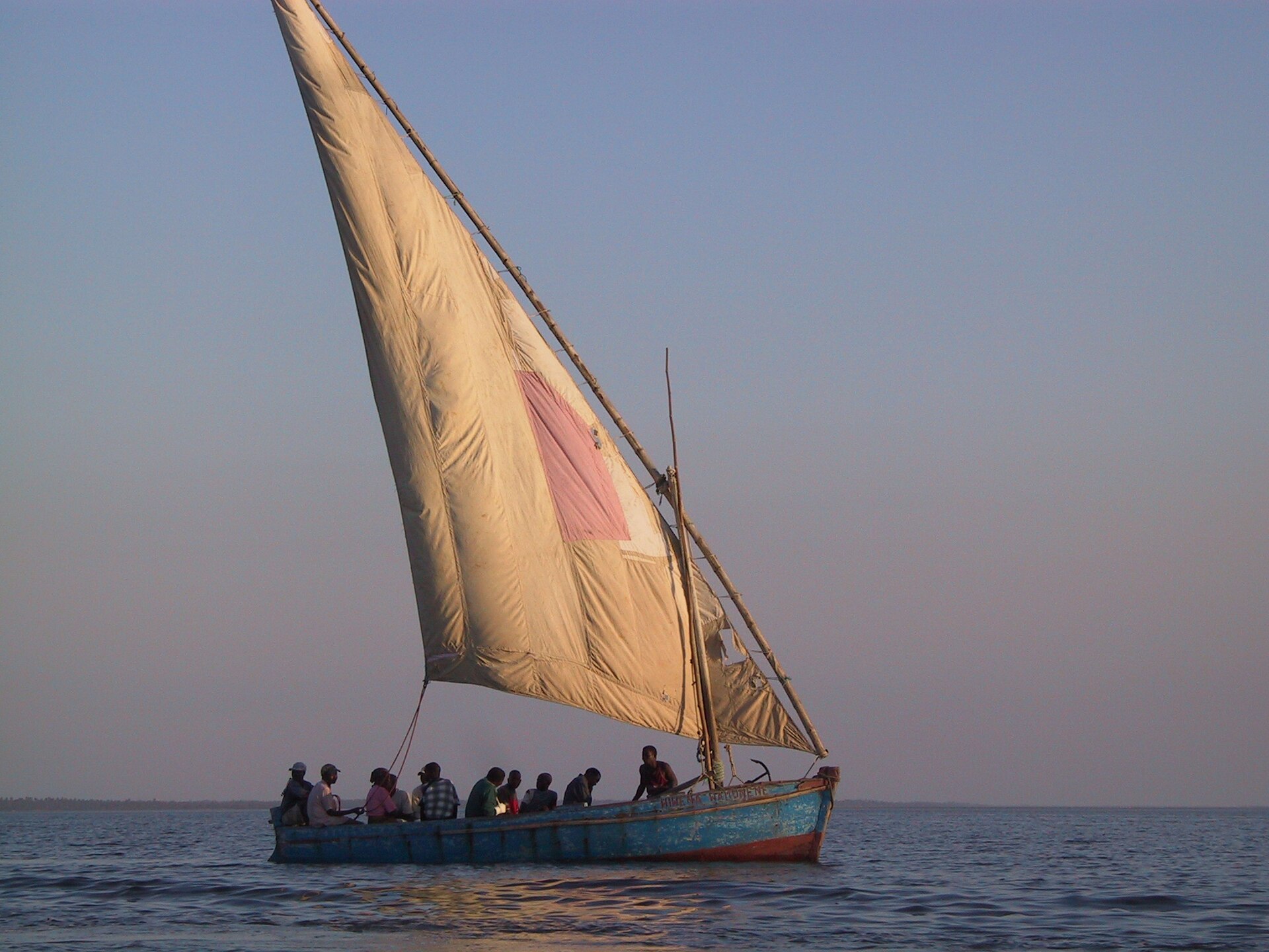 Dhow de casco azul descascado com uma grande vela bege remendada de rosa, levando cerca de dez pessoas no mar calmo ao entardecer