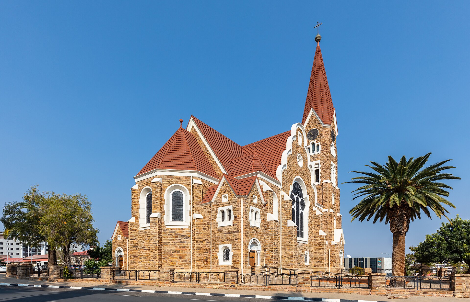 Christuskirche, igreja de arenito com telhado vermelho e torre com relógio, vista de lado sob céu azul, com uma palmeira à direita
