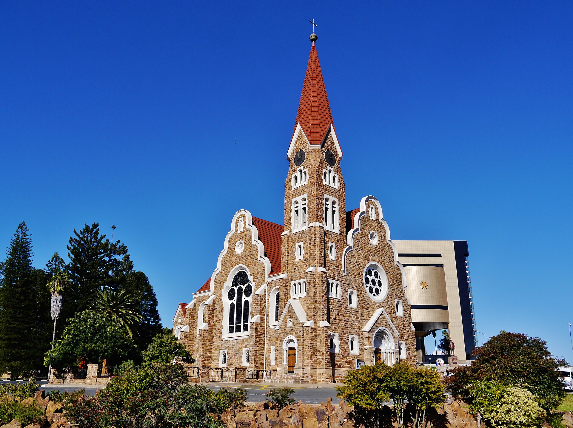 Christuskirche em arenito com torre de telhado vermelho e, à direita, o prédio dourado e escuro do Independence Memorial Museum, sob céu azul intenso