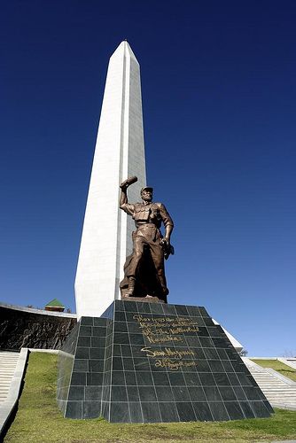 Obelisco branco atrás de uma estátua de bronze de soldado com o braço erguido, sobre pedestal de pedra escura com inscrições douradas, no Heroes' Acre