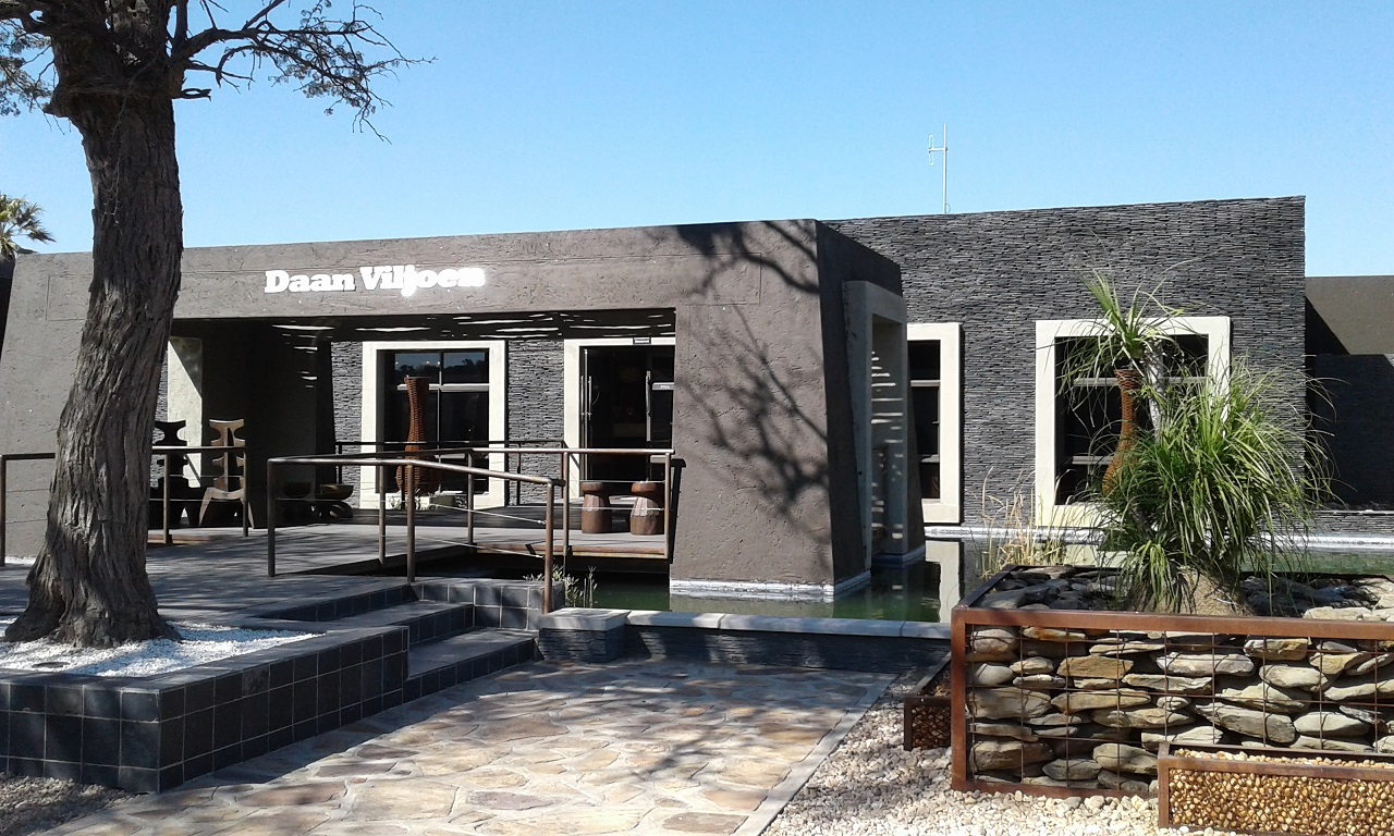 Entrada do Daan Viljoen Game Park, prédio de paredes escuras com o letreiro Daan Viljoen, espelho d'água, jardim de pedras e uma árvore à esquerda