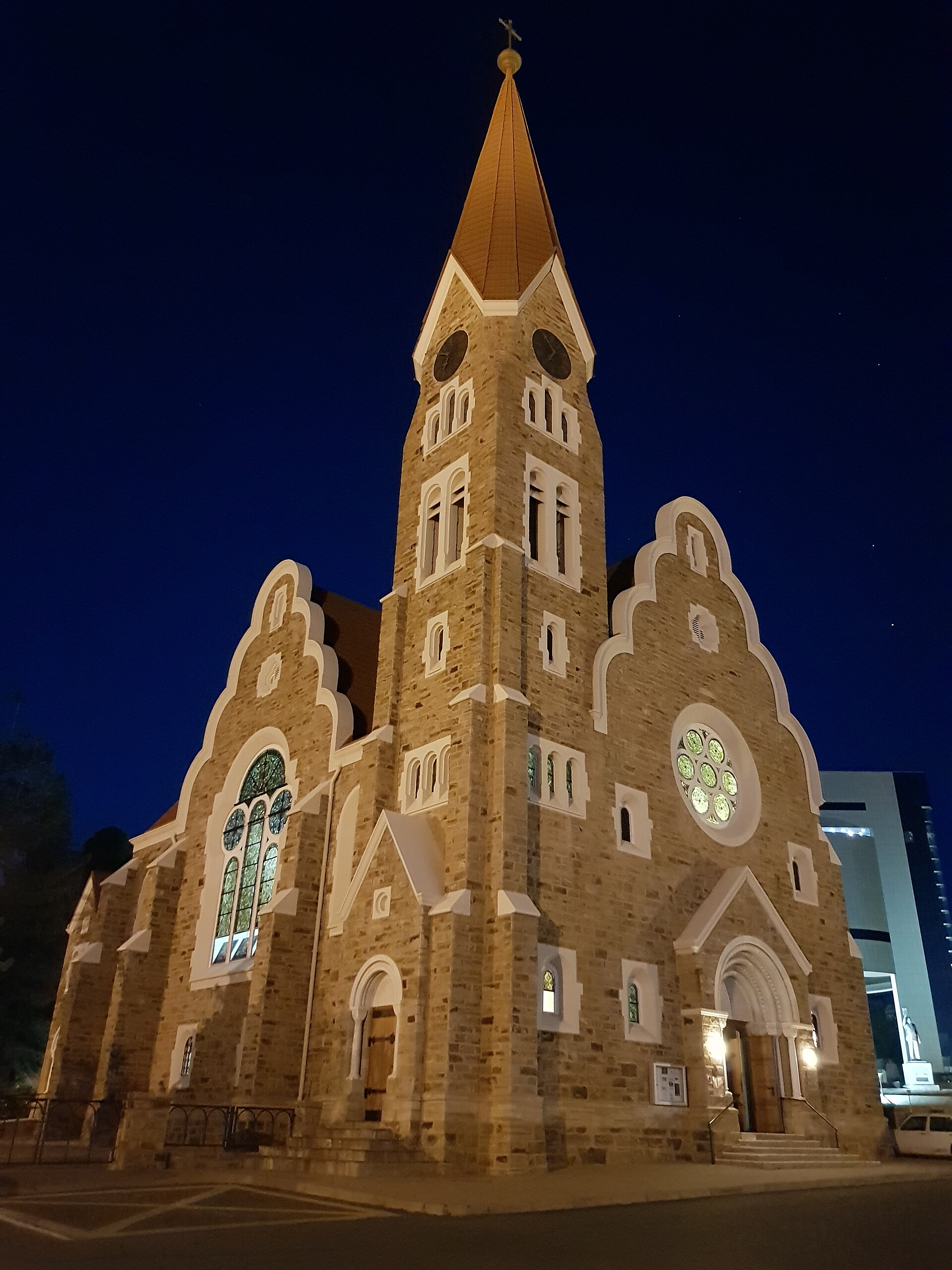 Igreja de pedra com torre alta e telhado pontiagudo iluminada à noite, com vitrais acesos e céu azul-marinho