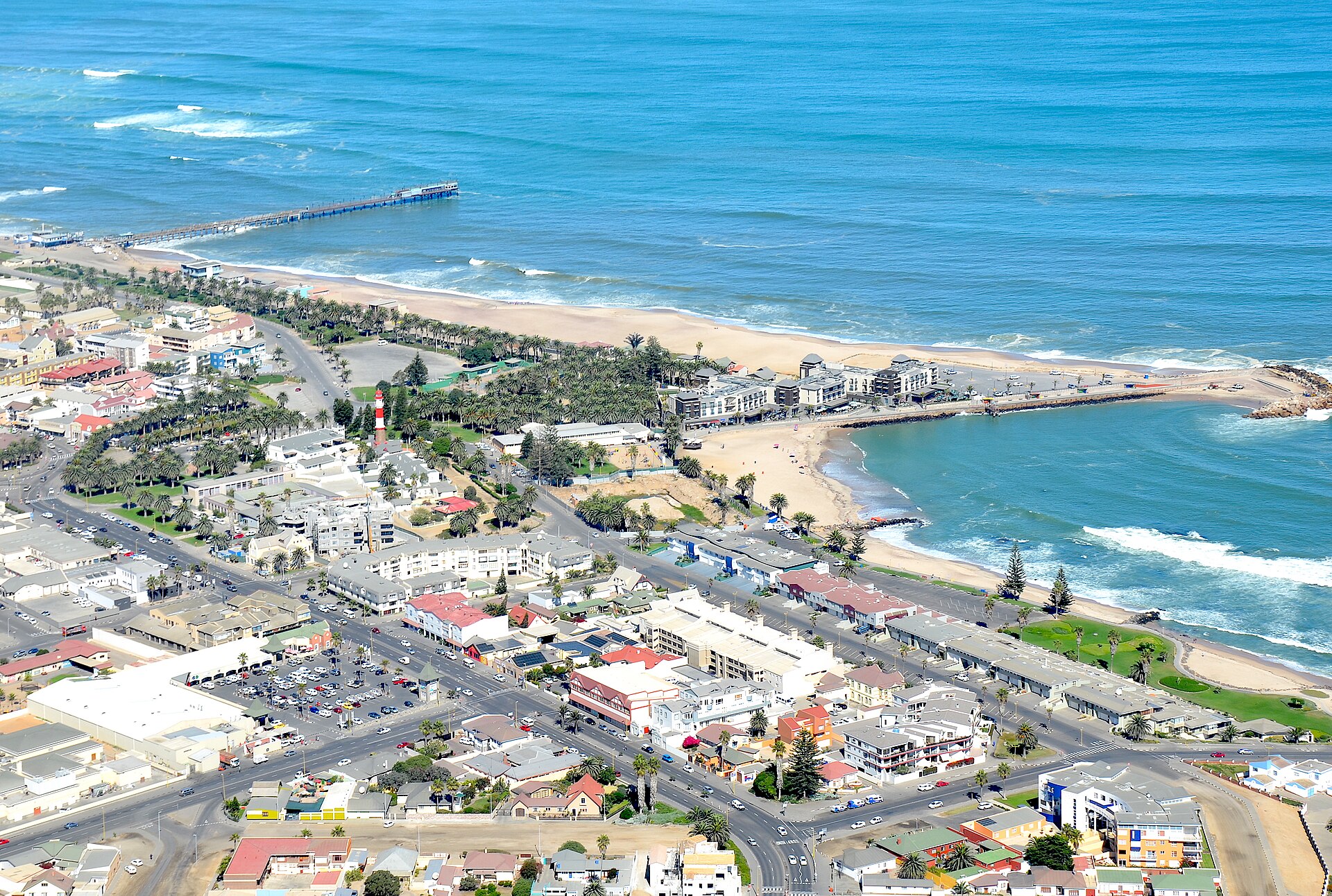 Vista aérea de Swakopmund, com ruas, palmeiras e prédios junto à praia, um píer comprido avançando no mar e um farol vermelho e branco