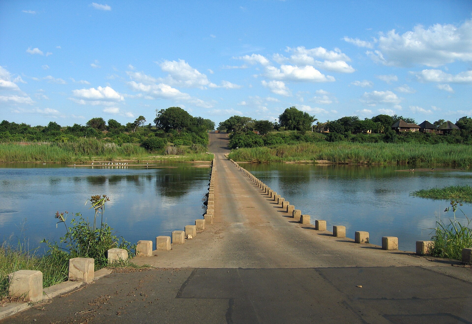 Ponte baixa de concreto com marcos de pedra nas bordas atravessando um rio calmo, com vegetação verde e chalés de telhado de palha na margem direita