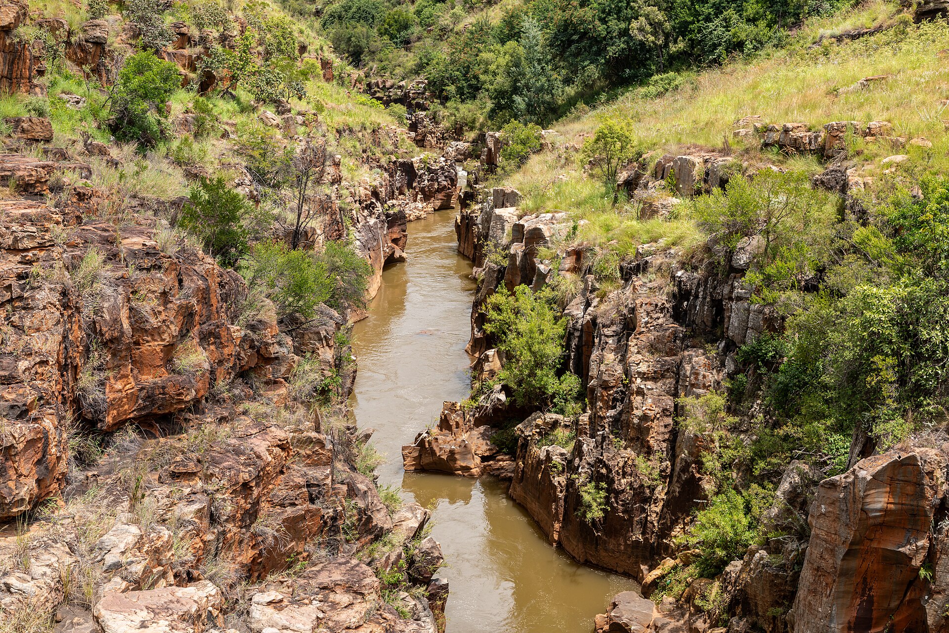Rio de água barrenta correndo por um desfiladeiro estreito de rochas alaranjadas, com mato verde e capim nas encostas
