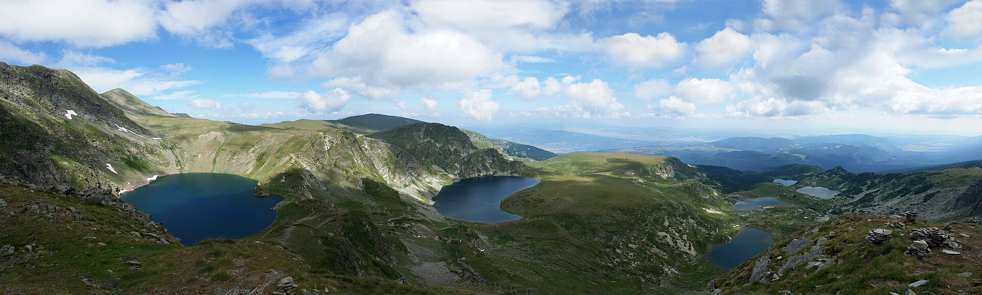 Panorama de montanha com três lagos escuros entre encostas verdes e manchas de neve, sob céu azul com nuvens