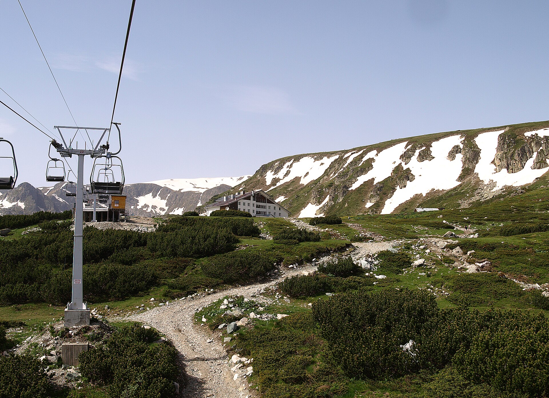 Cadeiras do teleférico ao lado de um poste, com uma cabana de montanha e uma trilha de pedra entre arbustos e encostas com neve