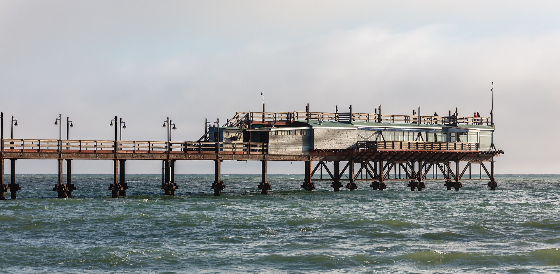 Píer de Swakopmund sobre estacas de ferro enferrujado, com um restaurante de paredes cinzentas no fim da passarela e o mar verde-acinzentado ao redor
